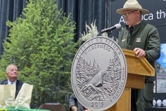 Garbed in a replica 1910 uniform, Charles Mark, Salmon-Challis National Forest supervisor, speaks as the Frank Church River of No Return Wilderness quarter is released on Wednesday, Nov. 6, 2019, during a ceremony at Salmon Junior-Senior High School in Salmon, Idaho. David Ryder, director of the U.S Mint, is seated behind currency bags at left, The coin is the 50th in the U.S. Mint’s America the Beautiful series of quarters. (© 2019 Cindi Christie/Cyanpixel)